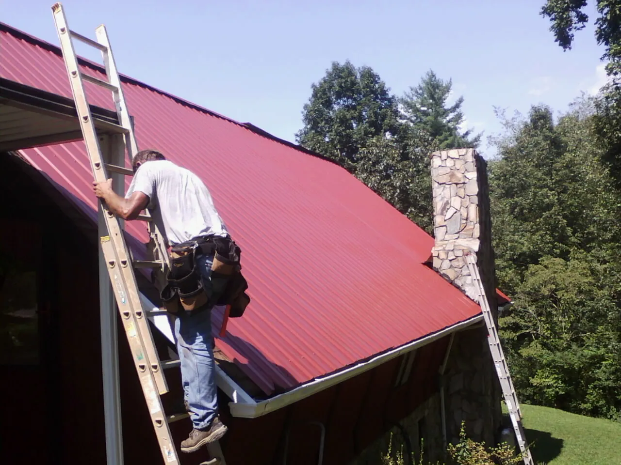 Roofer on a ladder beside a red metal roof and stone chimney — Soffit & Fascia Repair by Roofing Brothers in Clairton
